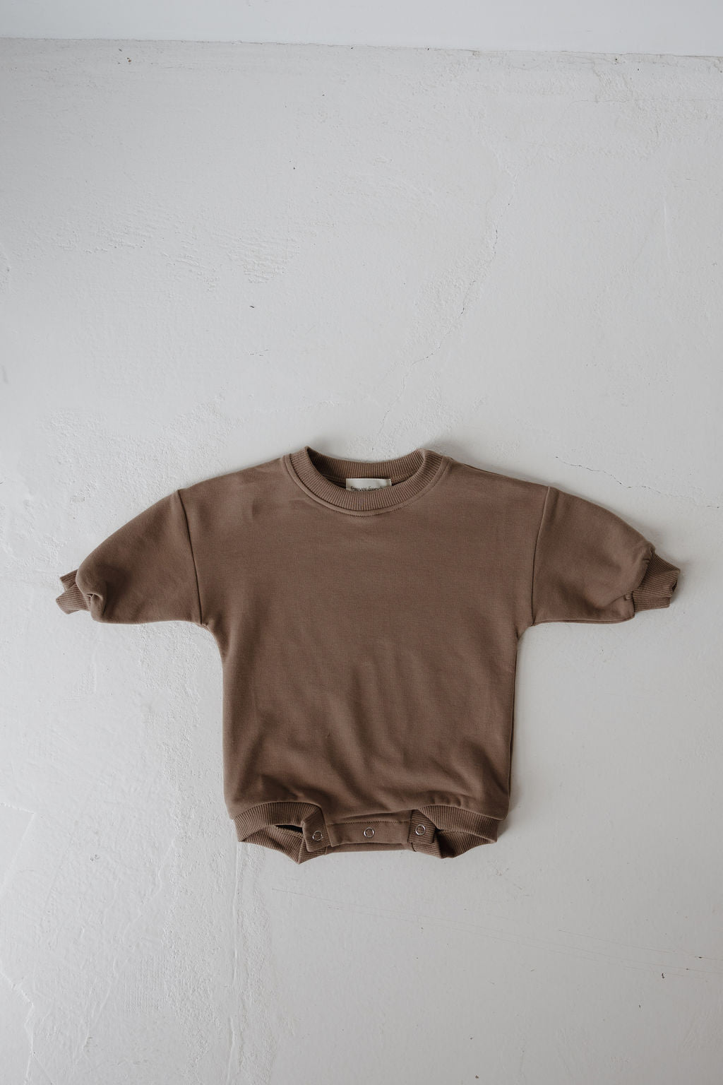 The forever french baby Sweatshirt Romper | Hot Cocoa is a brown long-sleeve baby onesie with rolled cuffs and a round neckline, laid flat on a white textured surface. Button snaps at the bottom make it perfect cozy wear for newborns.