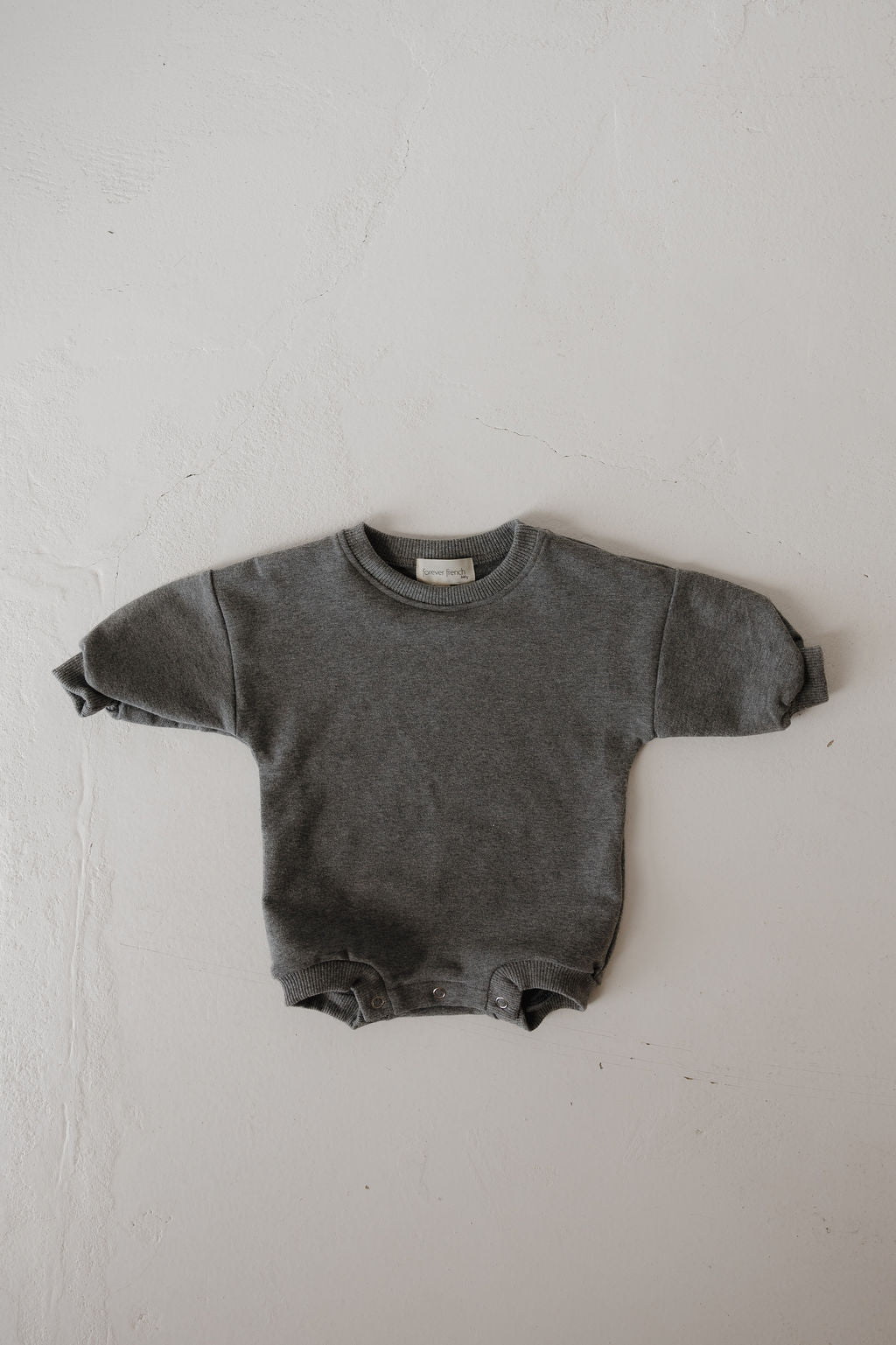 The Sweatshirt Romper | Charcoal by forever french baby is a gray long-sleeve romper with bottom snap buttons, laid flat on a light textured surface—ideal for newborns.