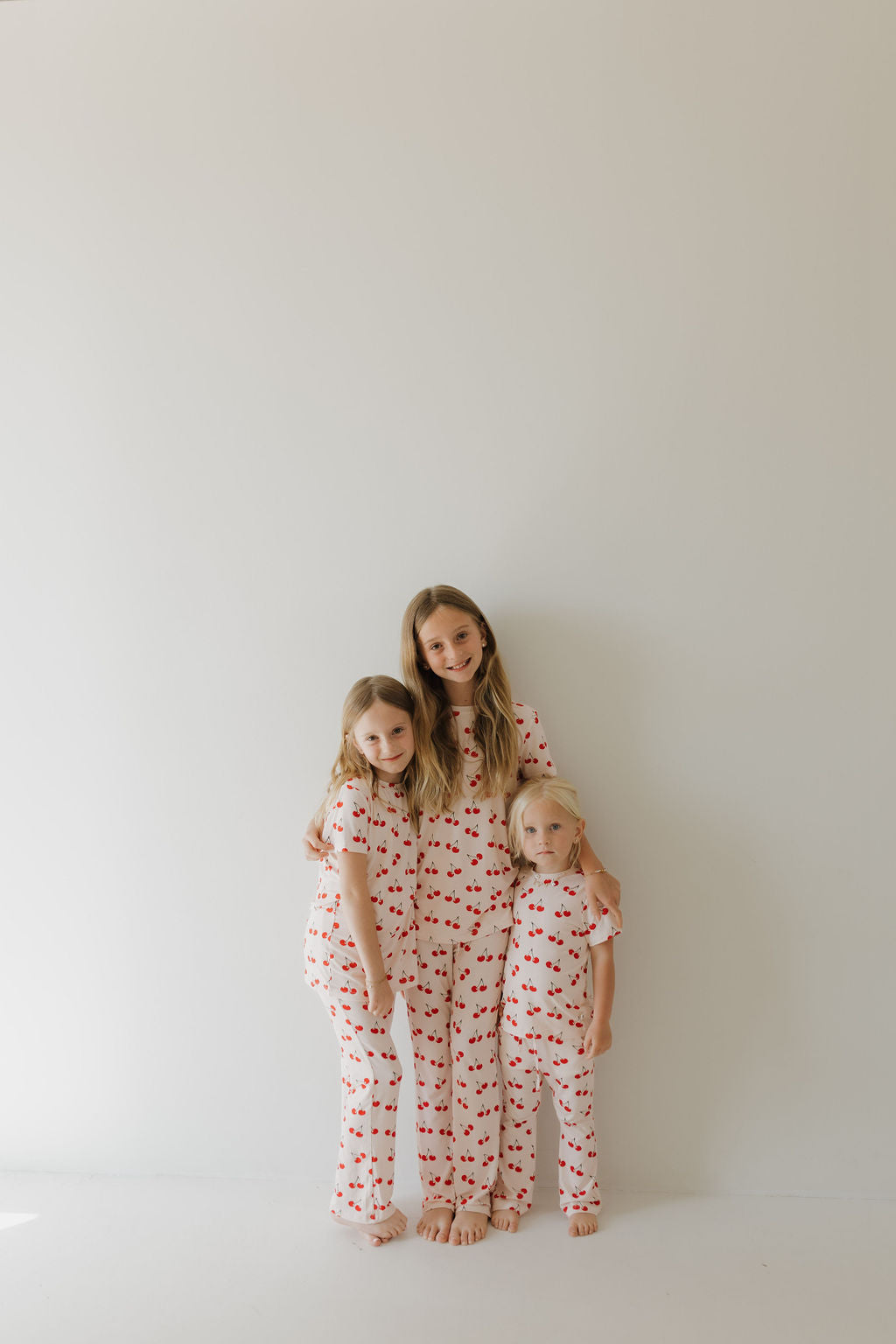 Three young children stand on a plain white background, wearing matching Toddler Short Sleeve Bamboo Lounge Sets in Cherry Orchard design by forever french baby. They are barefoot and smiling at the camera.