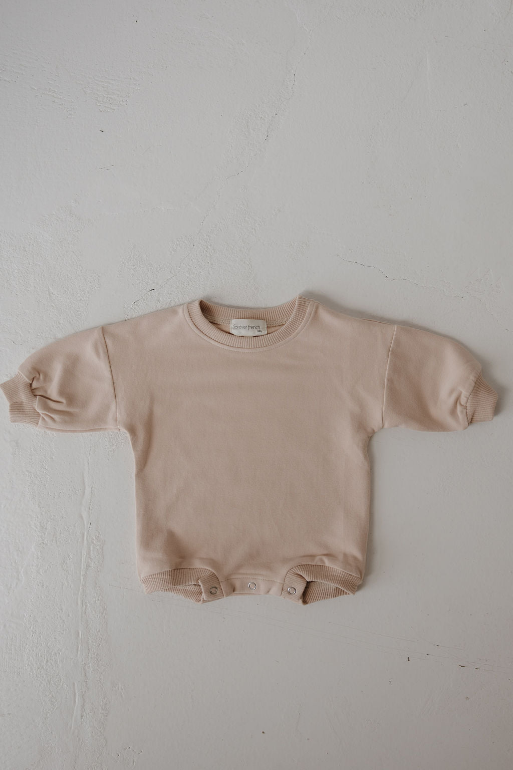 The forever french baby Sweatshirt Romper | Cashew is a beige long-sleeve romper with ribbed cuffs and snap buttons at the bottom, shown laid flat on a textured white surface.