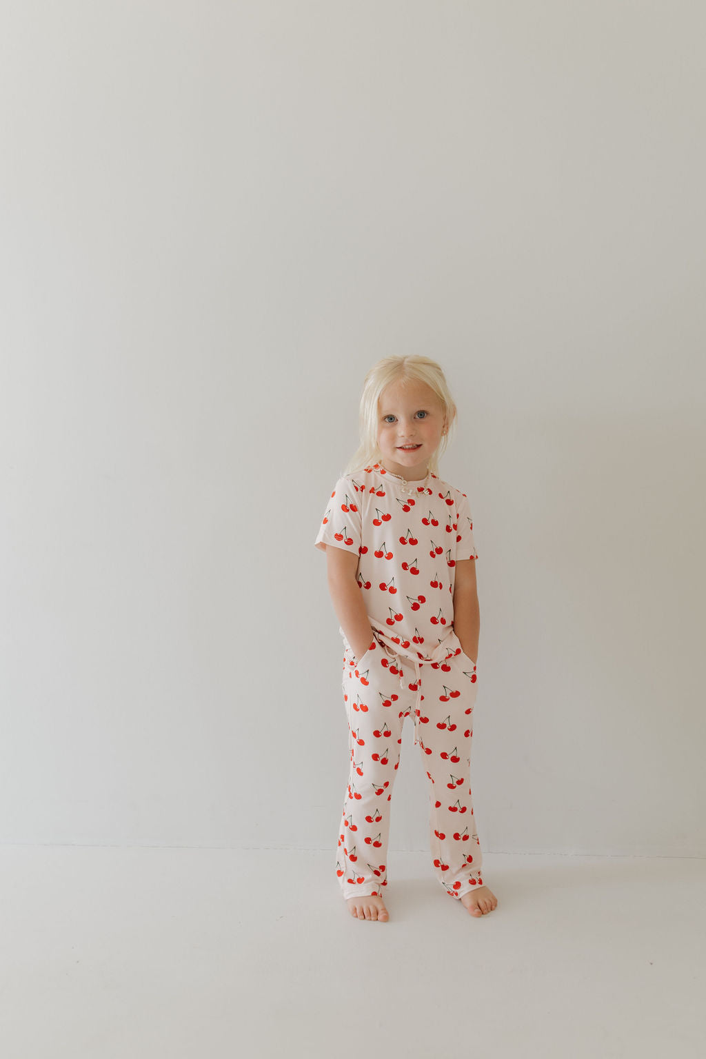 A young child with long blonde hair stands barefoot on a plain light background, wearing Forever French Baby's Toddler Short Sleeve Bamboo Lounge Set in Cherry Orchard. The breathable, hypo-allergenic material ensures comfort as they tuck their hands into pockets.