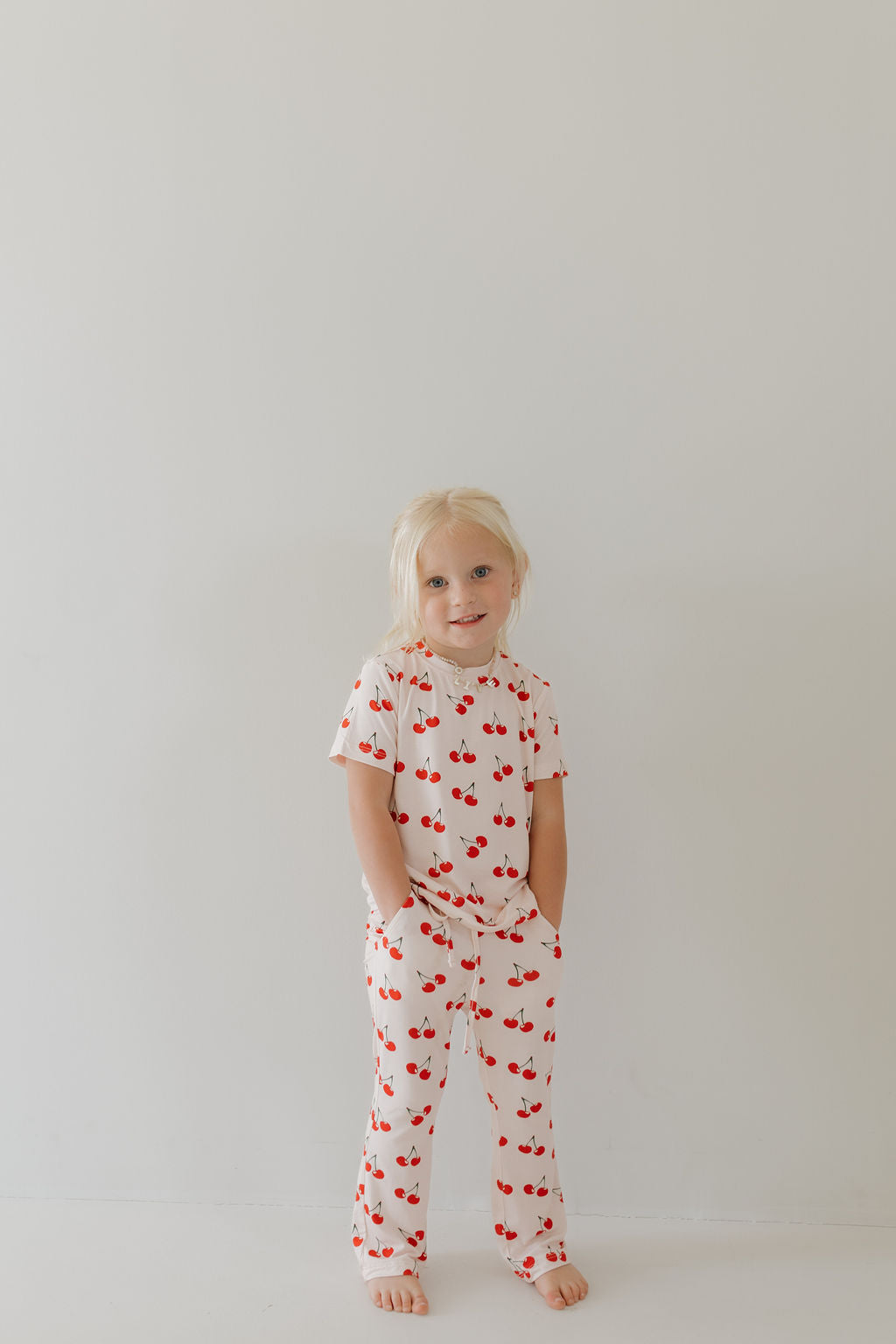 A blonde toddler stands barefoot, smiling against a white background in the forever french baby Toddler Short Sleeve Bamboo Lounge Set | Cherry Orchard. The hypoallergenic material ensures comfort for sensitive skin.