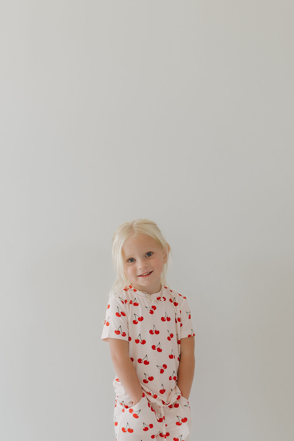 A young child with long blonde hair wears the hypo-allergenic "Toddler Short Sleeve Bamboo Lounge Set | Cherry Orchard" by forever french baby, featuring a white outfit with red cherry patterns. Crafted from breathable fabric, they stand against a light gray background, hands in pockets.