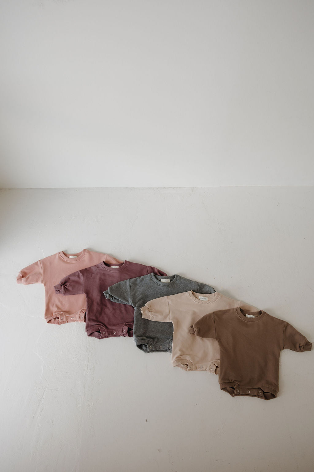 Five baby onesies in pink, maroon, gray, beige, and brown are lined up on a white floor—perfect for pairing with the forever french baby Sweatshirt Romper | Charcoal or other newborn outfits.