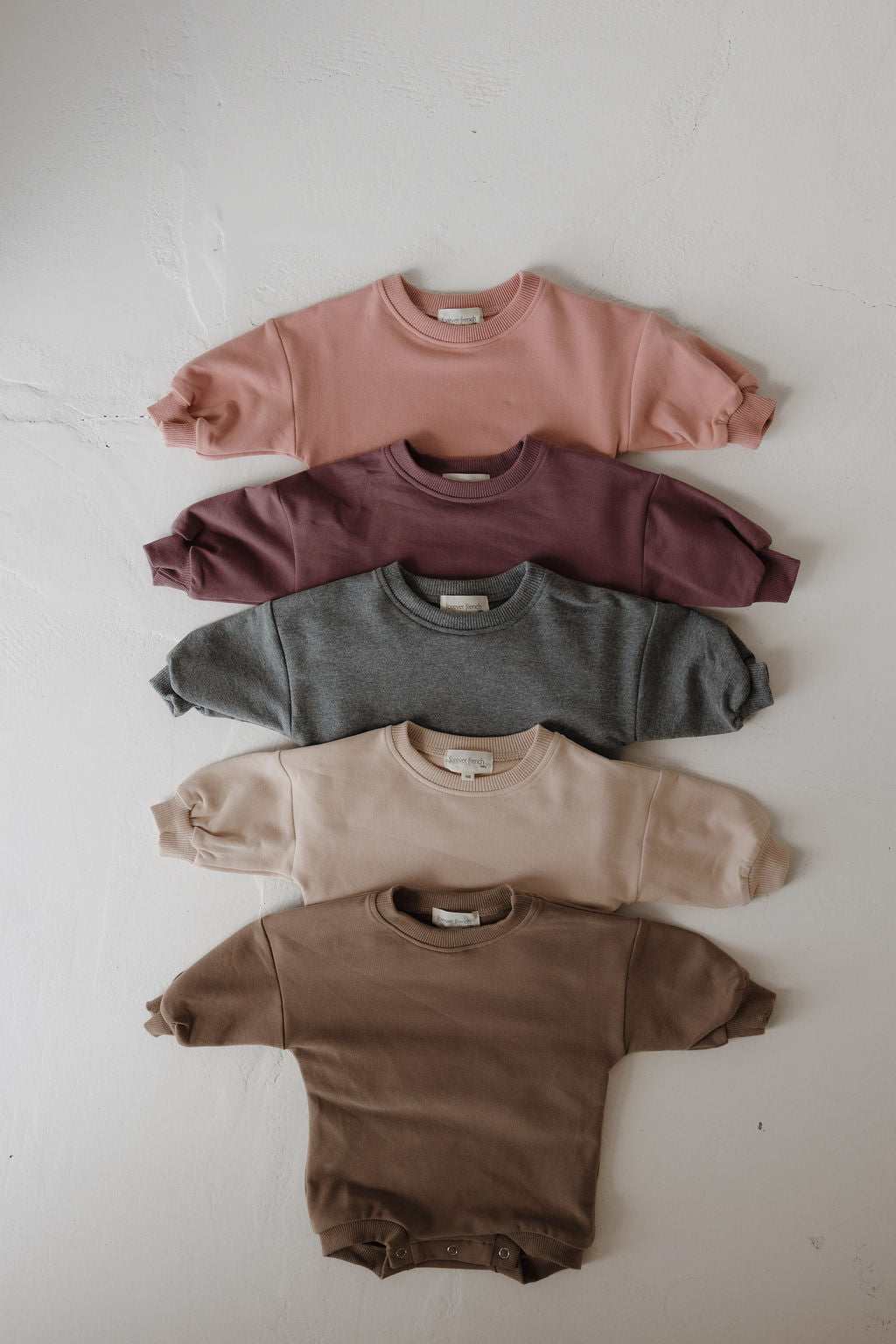 Five long-sleeve Sweatshirt Romper | Cashew by forever french baby in pink, purple, gray, beige, and brown are neatly stacked on a textured surface—perfect for pairing with a sweat set or as a cozy newborn romper.