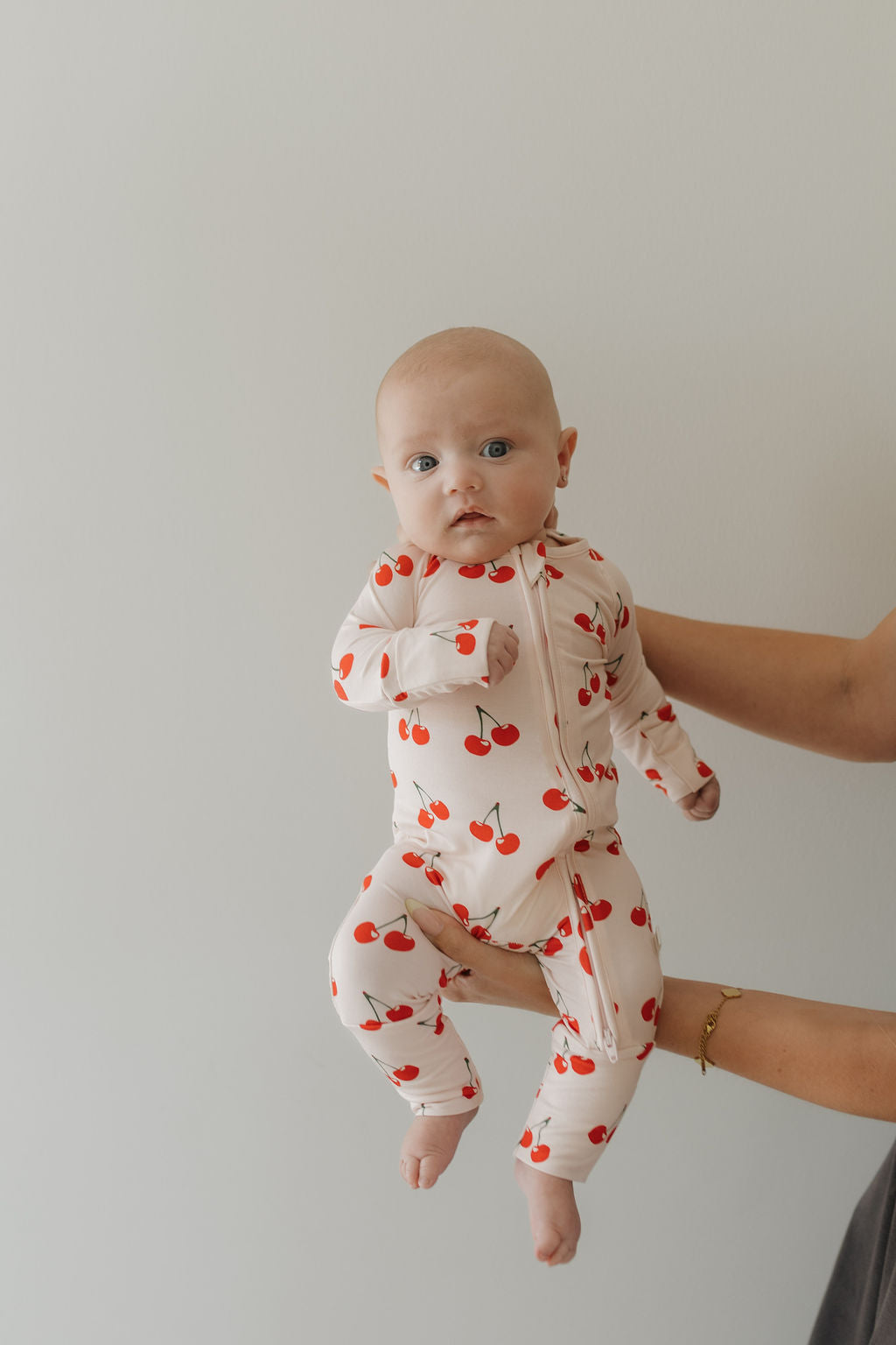 A baby in forever french baby's Bamboo Zip Pajamas | Cherry Orchard is held against a light background. The infant, wearing breathable, hypo-allergenic clothing, has wide blue eyes and looks at the camera while a hand offers gentle support from the side.