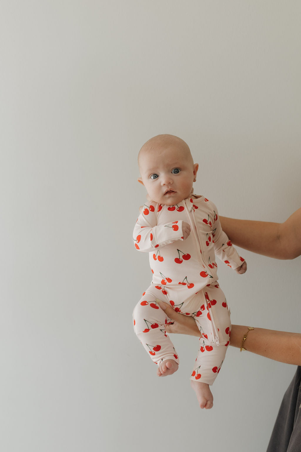 A baby in a Bamboo Zip Pajamas | Cherry Orchard by forever french baby is gently held against a plain light-colored wall. The baby gazes ahead, with only the holder's arm visible.