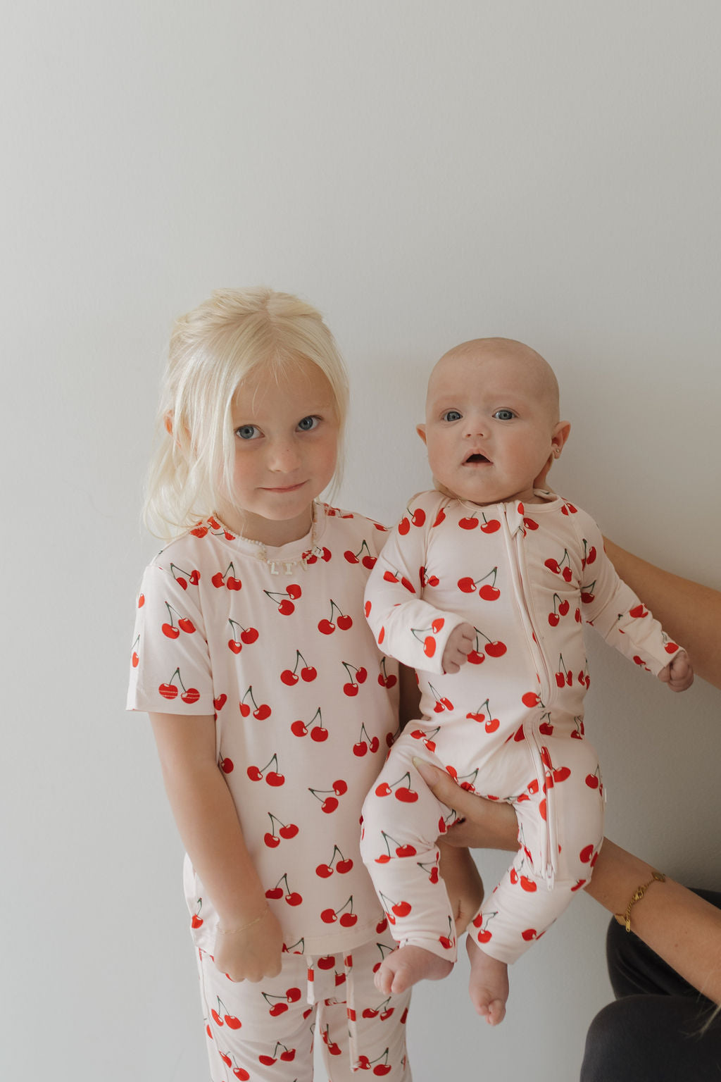 Two children in matching "Bamboo Zip Pajamas | Cherry Orchard" by forever french baby stand with an adult holding a baby in a cherry-patterned onesie. Set against a white background, their hypo-allergenic, breathable bamboo fabric attire shines with charm.