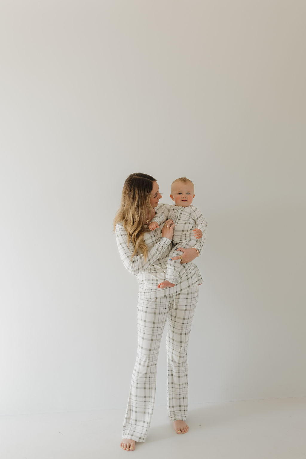 A woman in a forever french baby Women's Bamboo Pajama | Picnic Plaid cradles a baby wearing the same plaid pajamas, both made from breathable bamboo fabric. Standing barefoot on a plain background, the mother lovingly gazes at her child, while the baby looks directly at the camera.