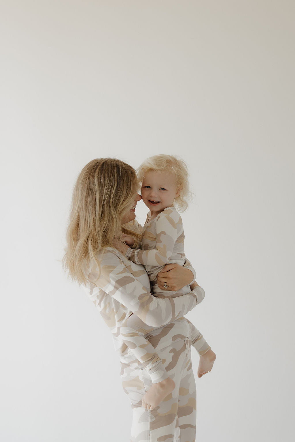 A woman with long blonde hair holds a smiling toddler as they wear matching forever french baby Women's Bamboo Pajamas | Camo 2.0, standing against a plain light background.