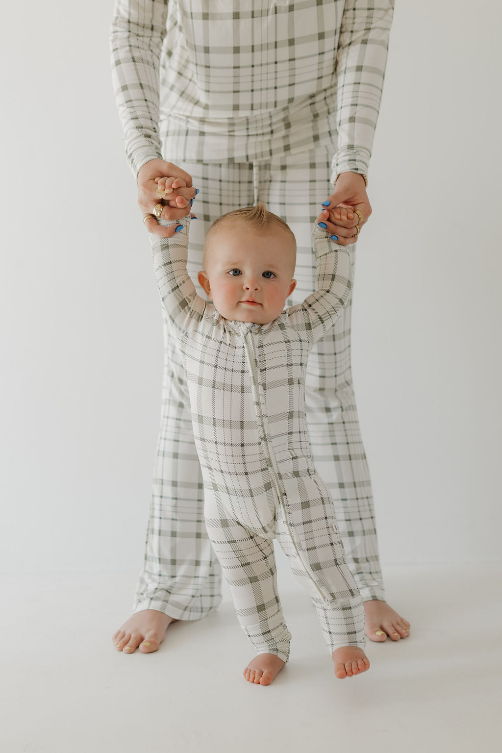 An adult, wearing the Women's Bamboo Pajama in Picnic Plaid by forever french baby, holds a baby's hands as they stand on a white background. The baby, with a small mohawk, also dons matching plaid pajamas.