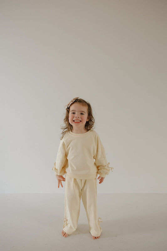 A young child stands barefoot and smiling in forever french baby’s Child's Sweat Set | Buttercream Bows, featuring a cream hue and bow details. The cozy long-sleeve top and pants stand out against the simple, light background.