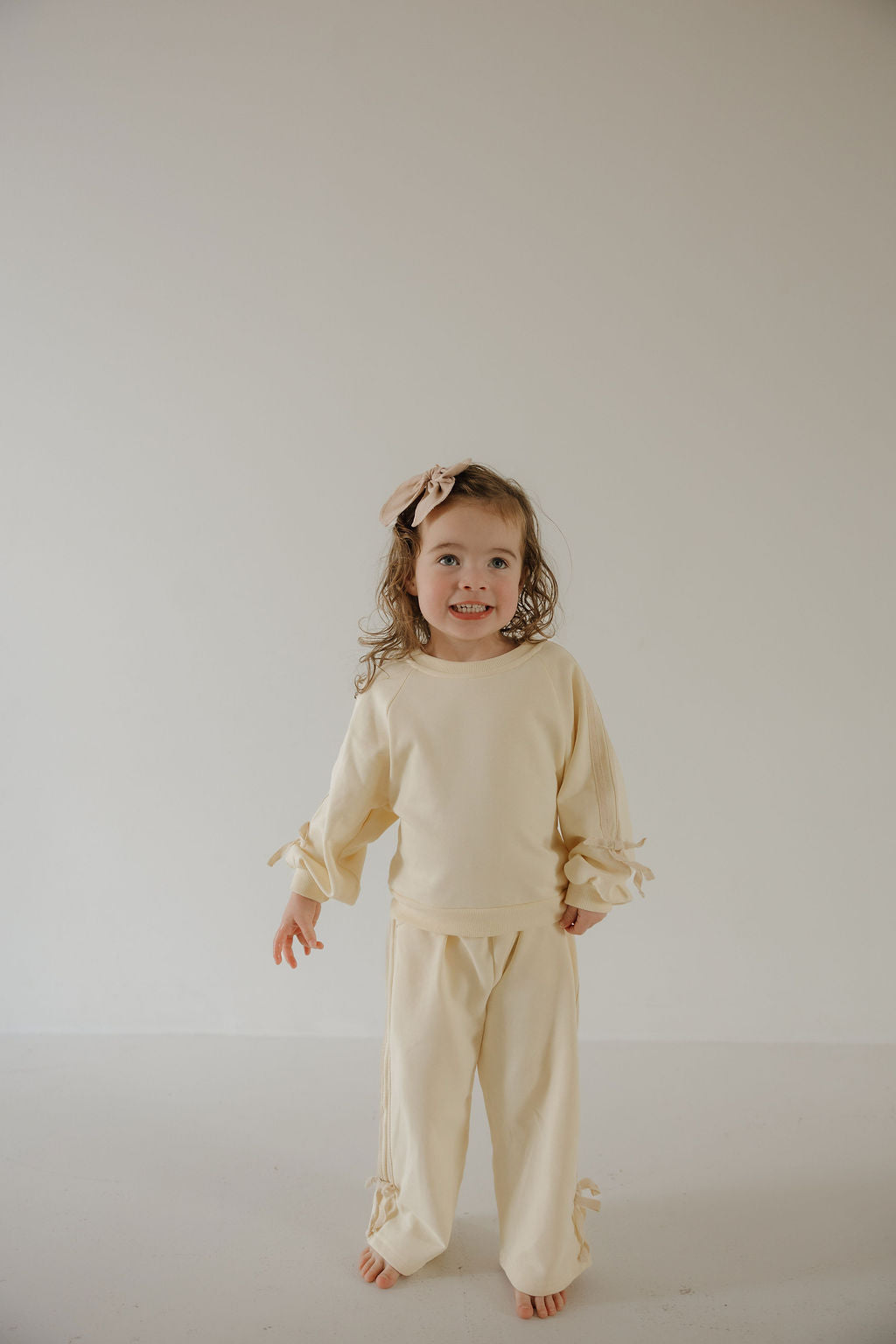 A young girl stands barefoot against a plain backdrop, smiling in the forever french baby Child's Sweat Set | Buttercream Bows, featuring bow details on the sleeves and pants and her shoulder-length curly hair loose.