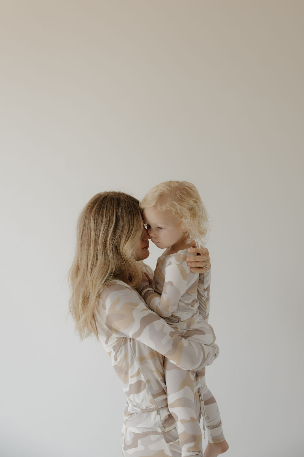 A woman with long blond hair holds a young child, both wearing forever french baby Women's Bamboo Pajama | Camo 2.0 sets. The breathable beige and white camo sleepwear contrasts against a plain, light background.