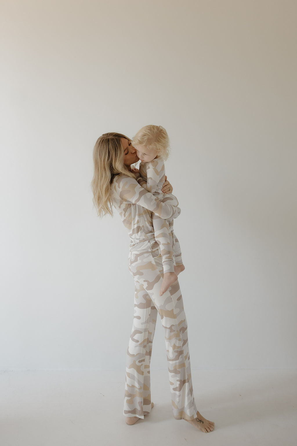 A woman in forever french baby Women's Bamboo Pajama | Camo 2.0 holds a young child, both in matching beige and white camouflage, standing barefoot as she kisses the child’s cheek. Their breathable sleepwear ensures comfort and softness.