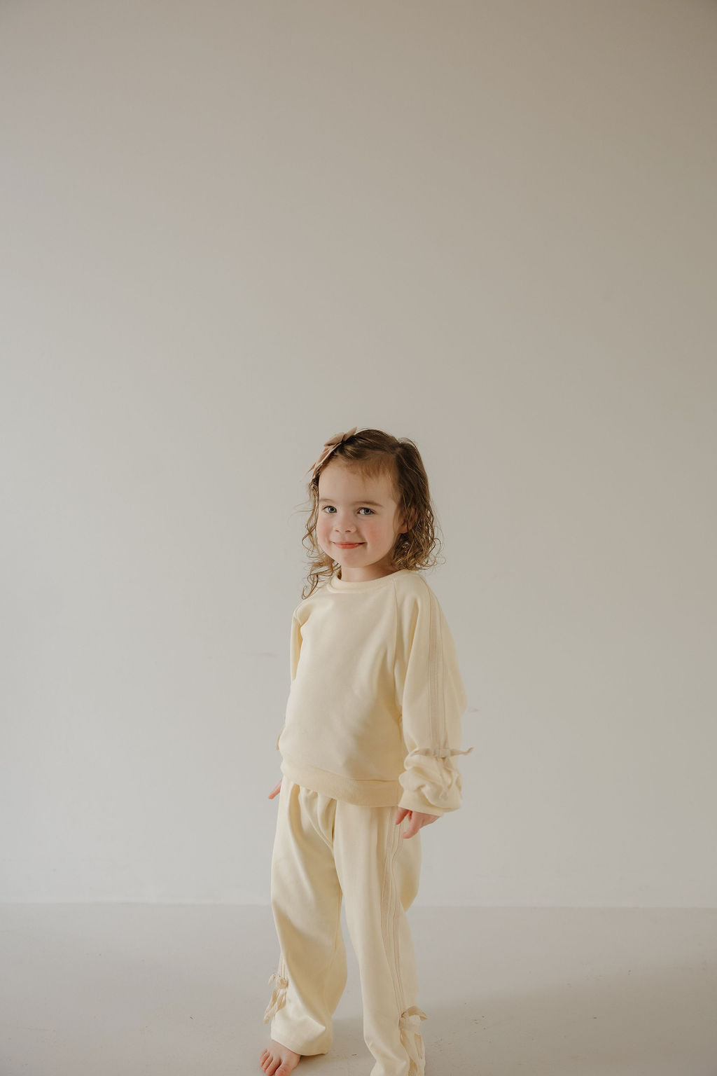 A young child with curly hair stands barefoot against a light background, wearing the forever french baby "Buttercream Bows" Child's Sweat Set featuring ruffle details on the sleeves and legs.