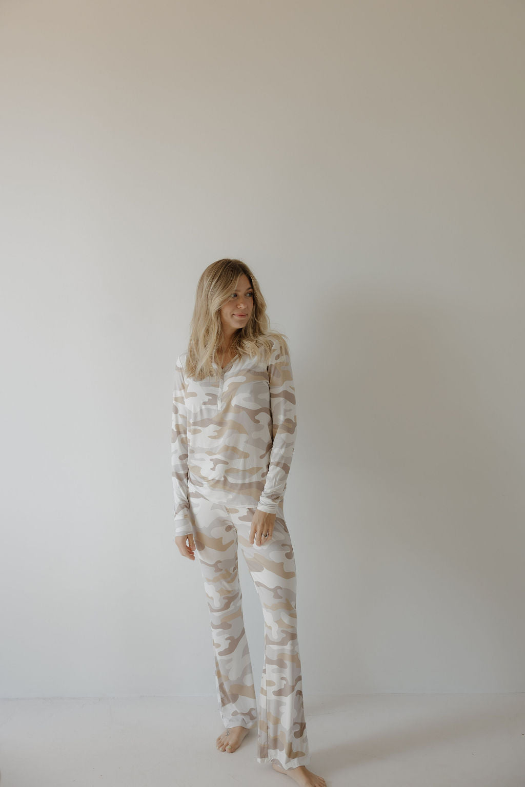 A blonde woman stands barefoot by a plain wall, wearing the forever french baby Women's Bamboo Pajama | Camo 2.0—a beige and white camo loungewear set with flared pants, crafted from breathable fabric for all-night comfort.