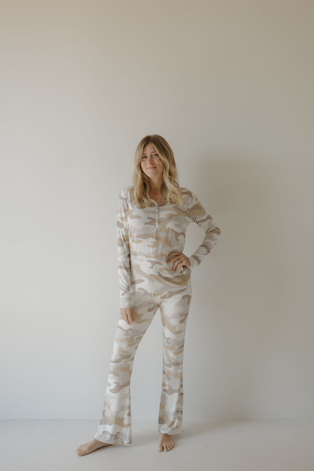 A woman with long blonde hair stands barefoot against a plain wall, smiling softly with one hand on her hip, wearing forever french baby Women's Bamboo Pajama | Camo 2.0—a hypo-allergenic beige and white camouflage-patterned set.