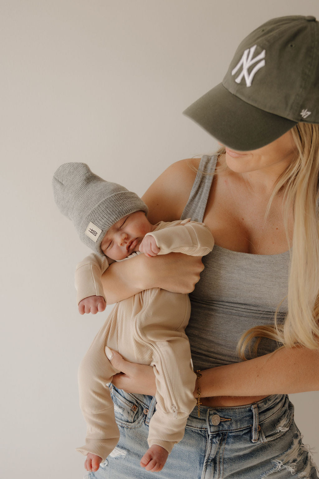 A woman in a gray tank top and NY cap holds a sleeping baby wearing forever french baby's Bamboo Zip Pajamas | Ribbed Cream and a gray knit hat, both softly lit to highlight the pajamas' premium bamboo comfort.