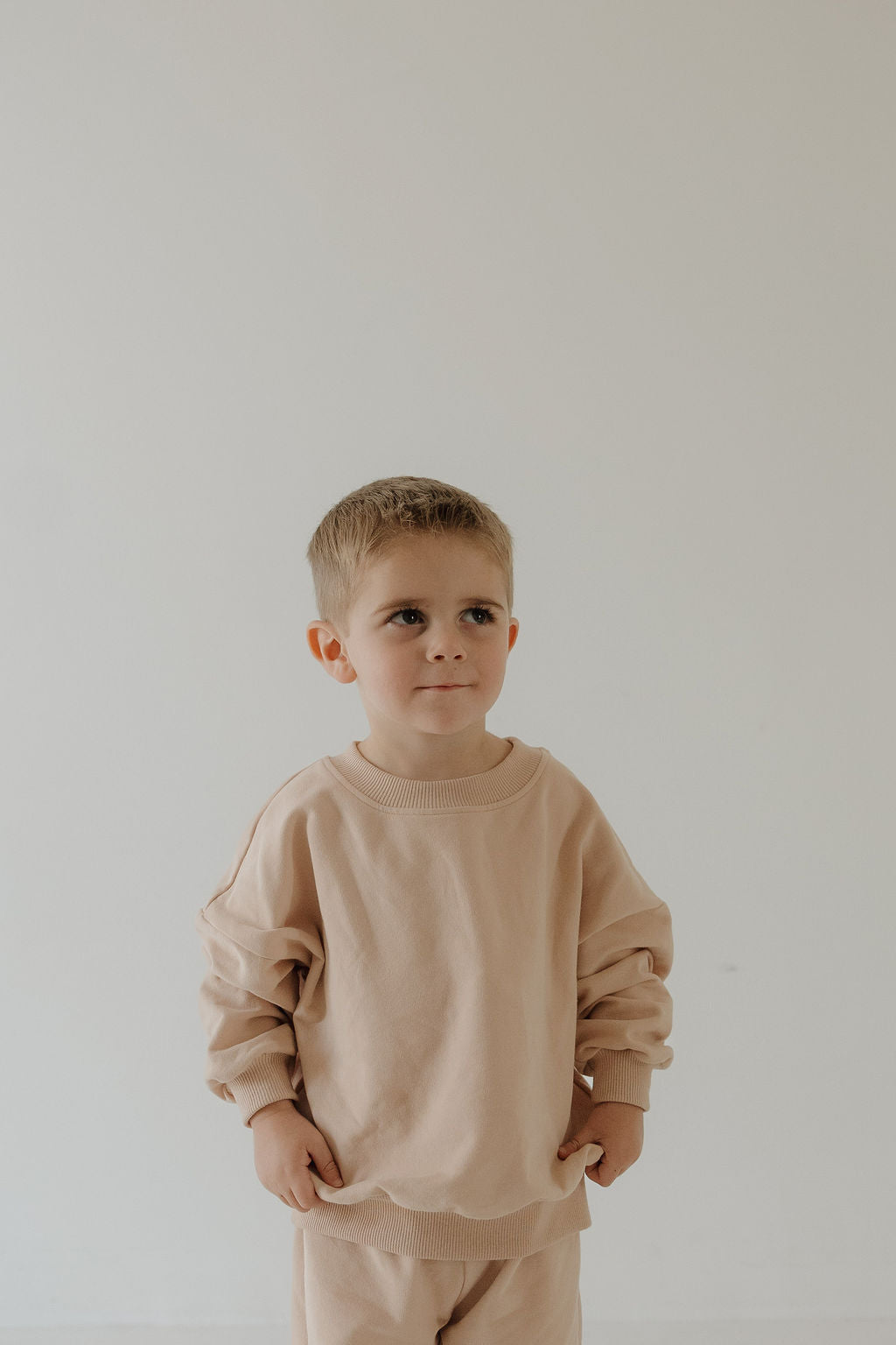 A young boy with short blond hair stands against a light background wearing the forever french baby Child Sweat Set in Cashew, an ideal beige outfit for everyday adventures, with his hands at his sides.