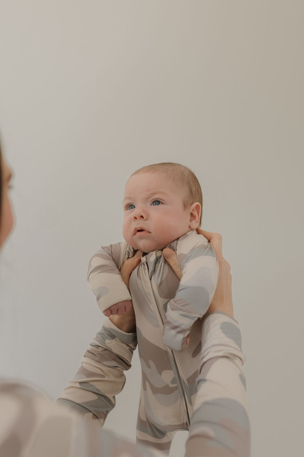 A person holds up a baby dressed in the Bamboo Zip Pajamas | Camo by forever french baby. The infant’s blue eyes gaze upwards, complementing the soft, neutral background.