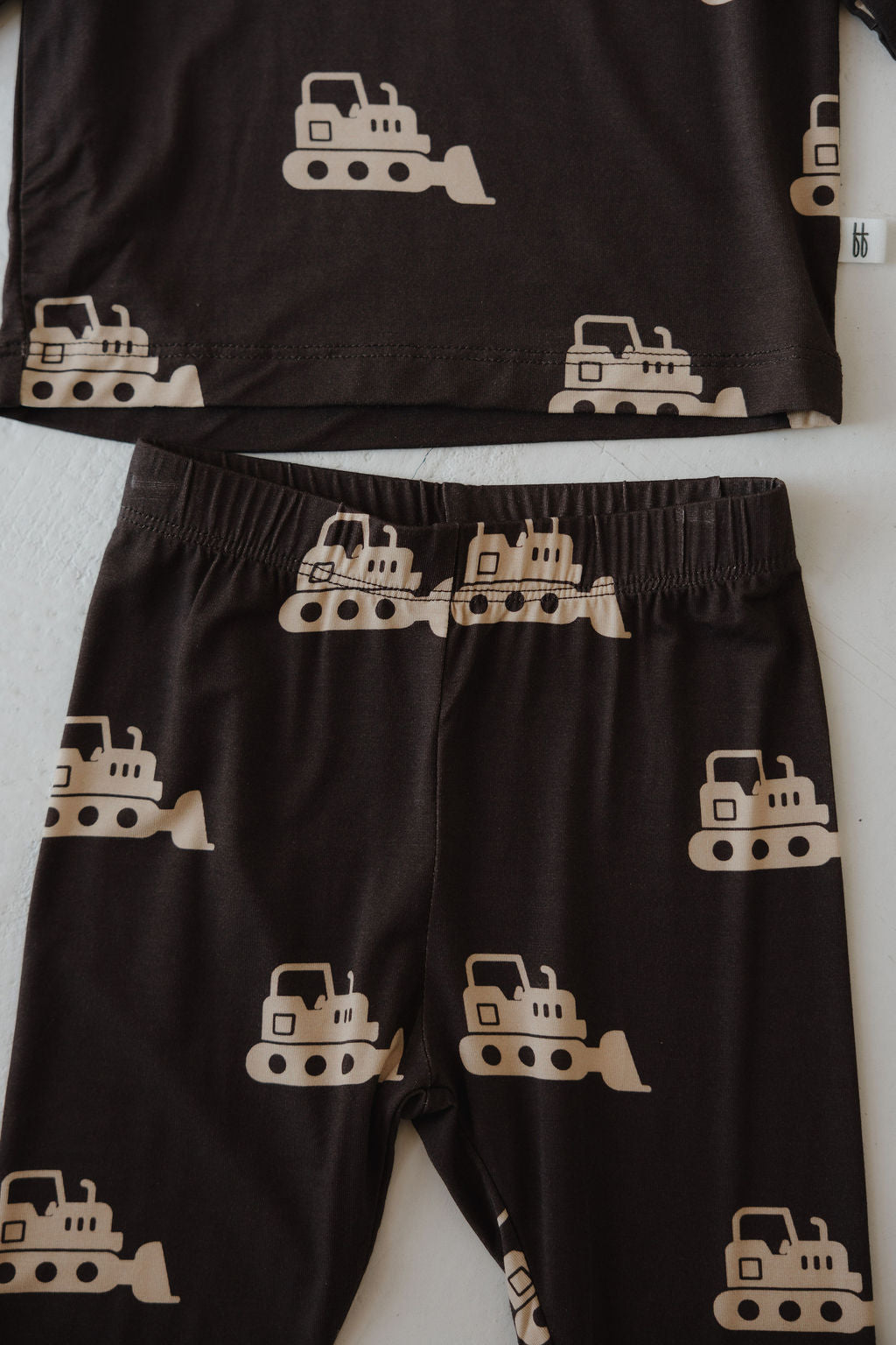 The forever french baby Child Sleepy Time Set | Bulldozer is a black bamboo outfit for kids, featuring hypo-allergenic fabric and a beige bulldozer print. The set includes both a top and pants with repeating bulldozer illustrations.