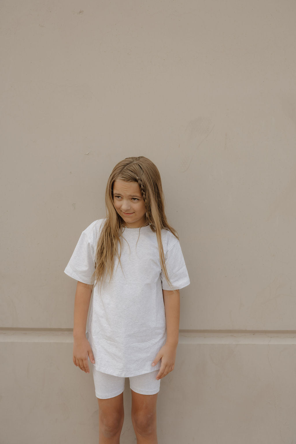 A young girl with long, light brown hair stands against a beige wall, wearing the forever french baby Child Short Set in Heather, gazing slightly to her left with a neutral expression.