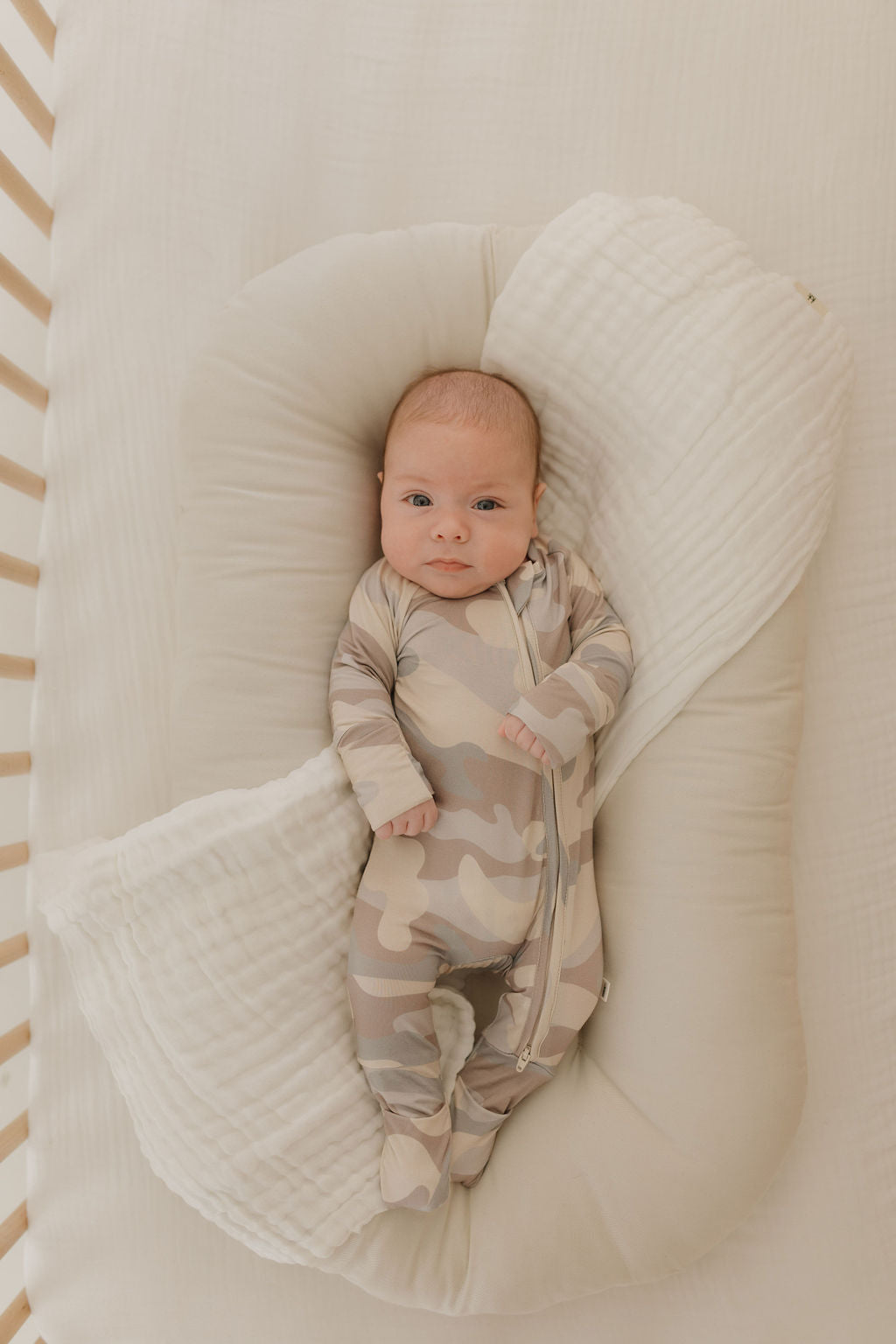 A baby wearing forever french baby Bamboo Zip Pajamas in camo rests on a soft, white baby nest in a crib. The hypo-allergenic fabric ensures comfort while the awake baby gazes up with a calm expression.