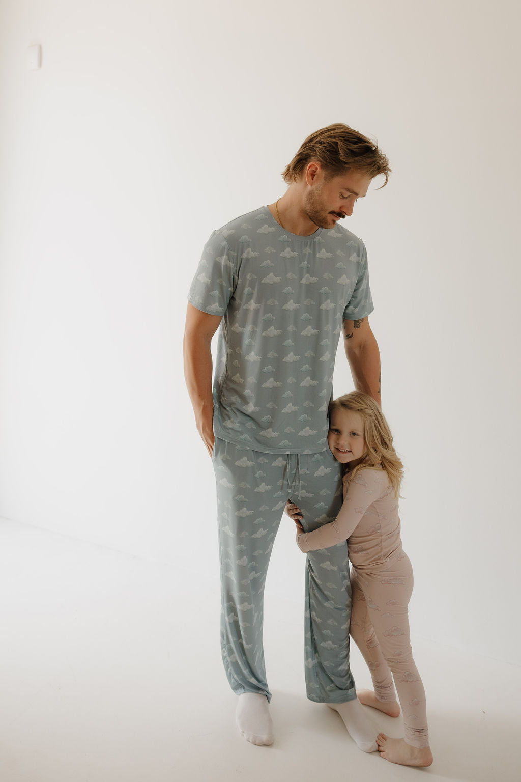 A man wears Kendy 3's Bamboo Two Piece Pajamas | Ruby in light blue with clouds, standing barefoot in a bright room as a girl in light pink pajamas hugs his leg, both enjoying the soft, breathable comfort.