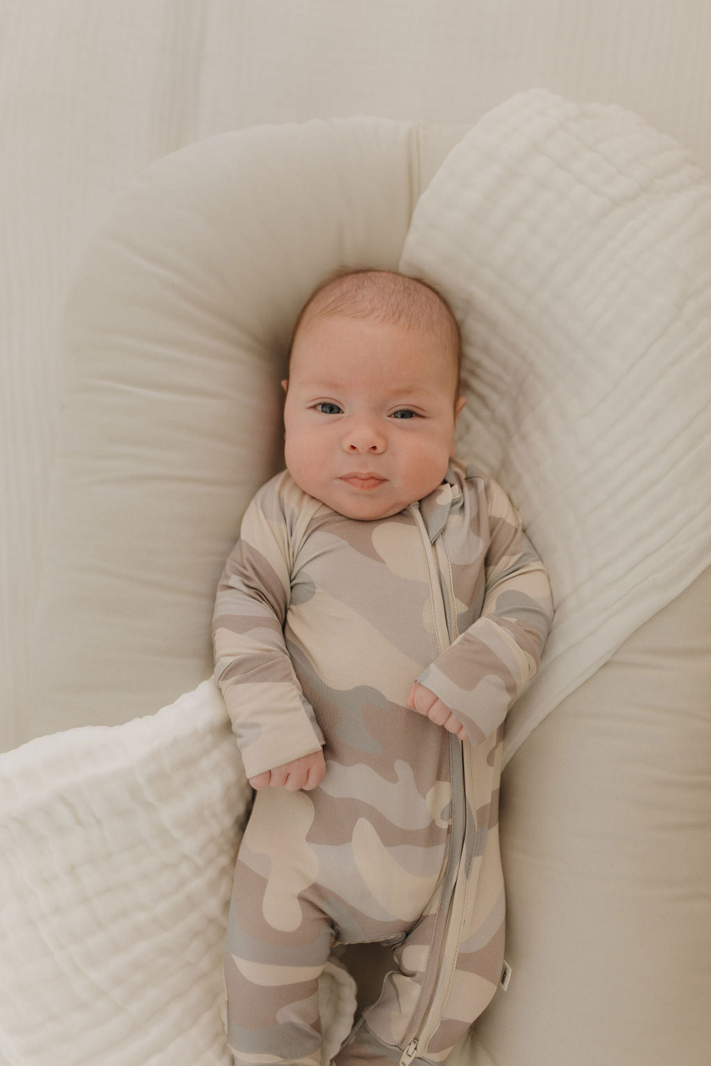 A baby, comfortably dressed in a Camo Bamboo Zip Pajama from forever french baby, lies on a soft white pillow. The breathable sleepwear keeps the baby calm and relaxed in a cozy, neutral-toned setting.