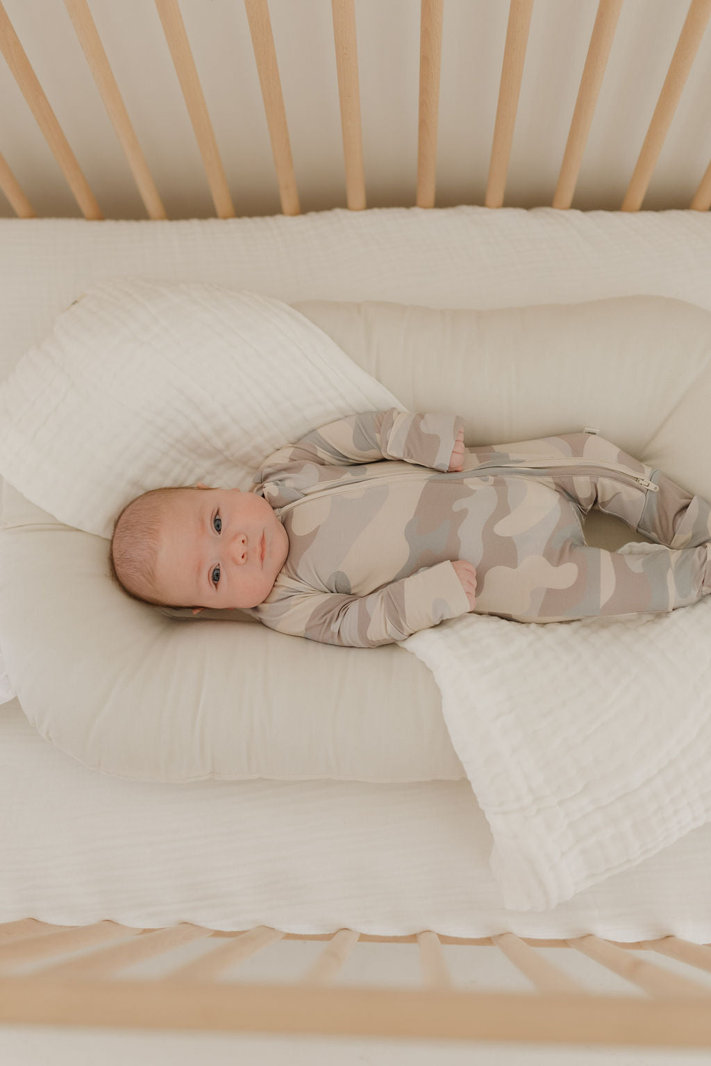 A baby wears forever french baby's Bamboo Zip Pajamas in a camo pattern, lying on a soft cream blanket in a crib. The hypo-allergenic wooden-slatted crib creates a calm, neutral-toned setting.