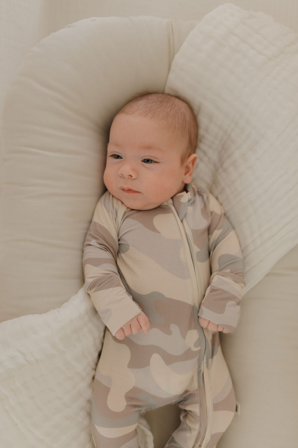 A baby rests on a soft surface in forever french baby's Bamboo Zip Pajamas | Camo. The breathable beige camouflage sleepwear ensures comfort as the child gazes to the side, surrounded by a textured, light-colored fabric background that enhances the serenity of the scene.