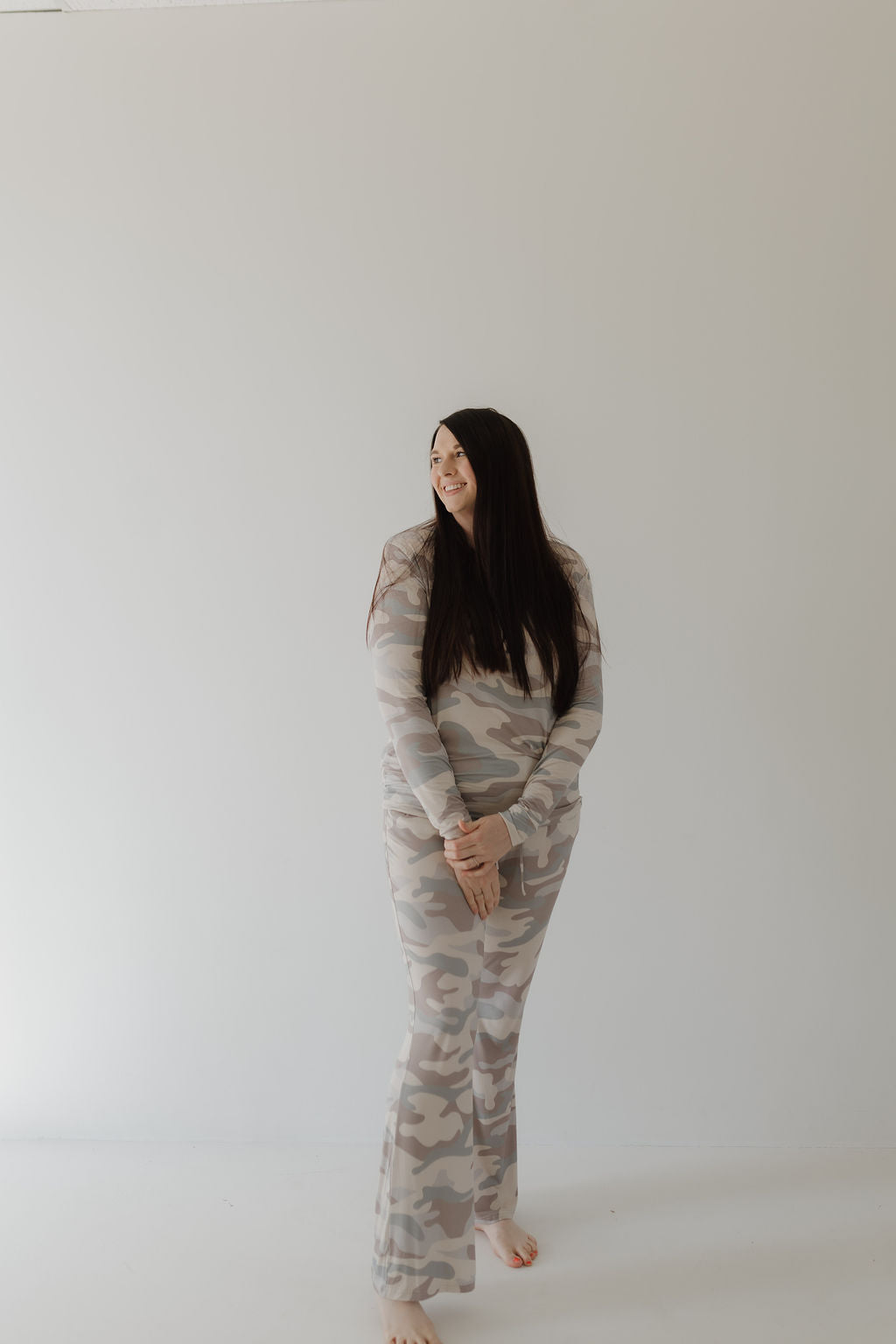 A woman with long dark hair stands barefoot against a plain background, wearing the forever french baby Women's Bamboo Pajama in a light camouflage pattern. The full-length, breastfeeding-friendly design complements her relaxed sideways glance.