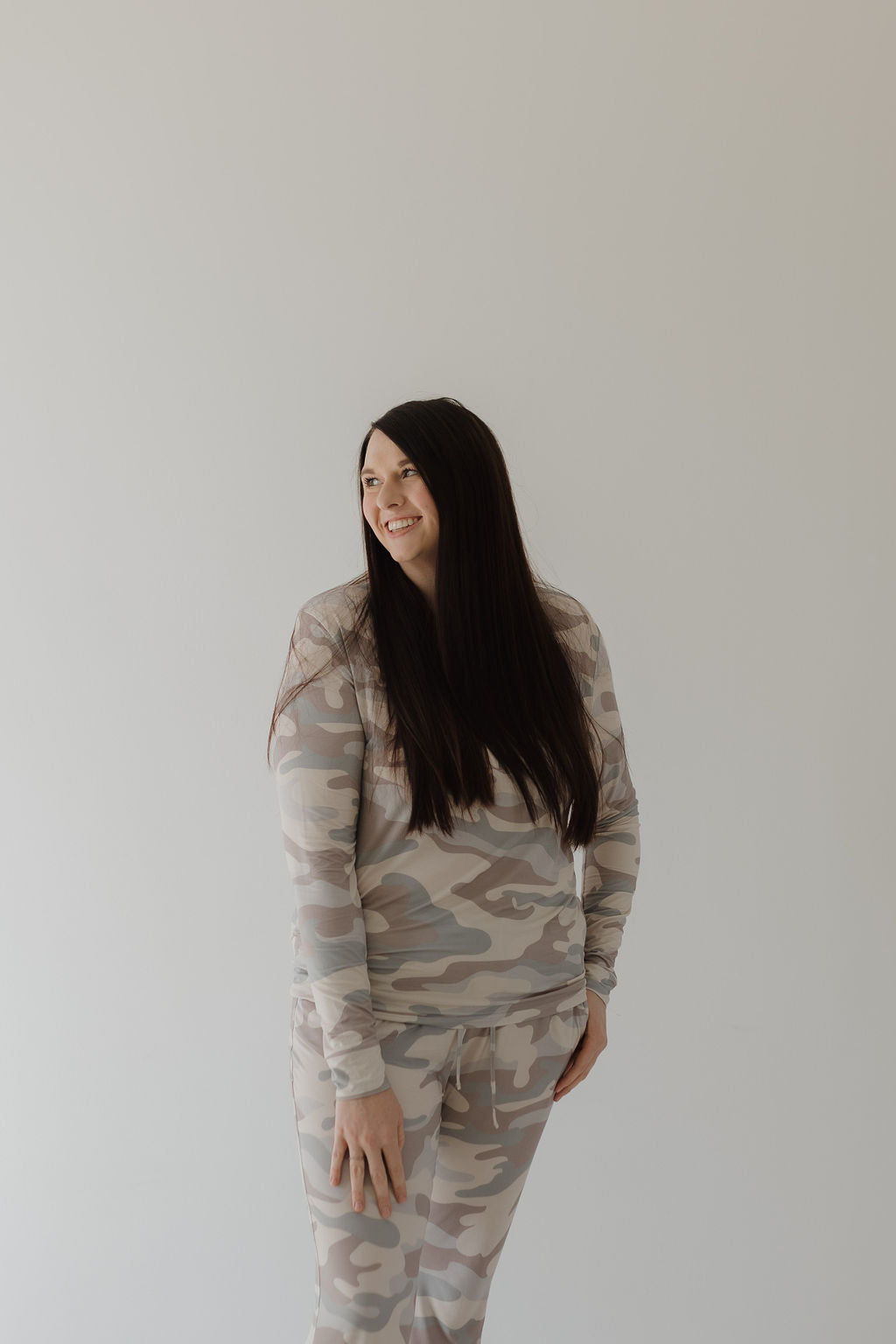 A woman with long dark hair, wearing "Women's Bamboo Pajama | Camo" by forever french baby, stands against a white background and smiles while looking left.