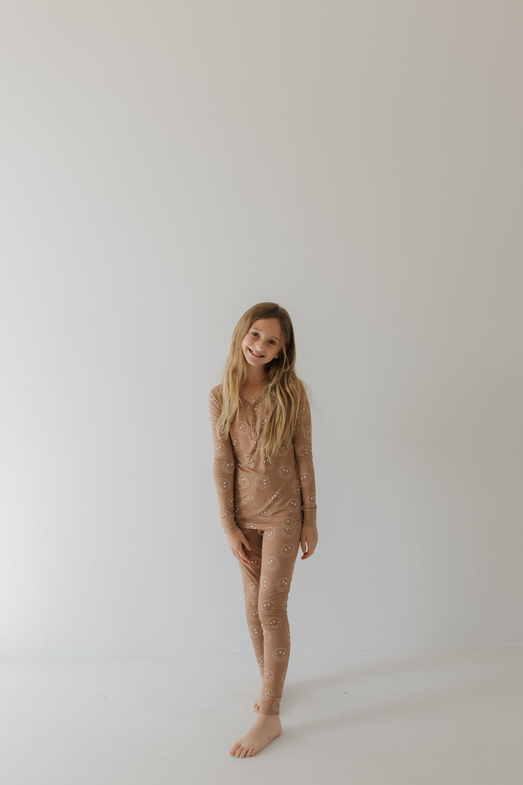 A young girl with long hair stands barefoot against a light background, smiling in a relaxed pose. She wears the "Bamboo Two Piece Pajamas | Sandstone Smile" by forever french baby, showcasing its breathable design with smiley face patterns.