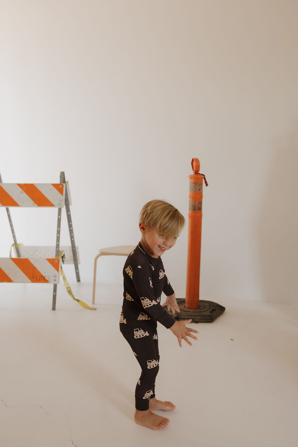 A young child smiles barefoot on a white floor wearing the forever french baby Child Sleepy Time Set | Bulldozer, featuring breathable pajamas with vehicle prints. Behind him are an orange traffic cone, barricade, and a chair against a white wall.