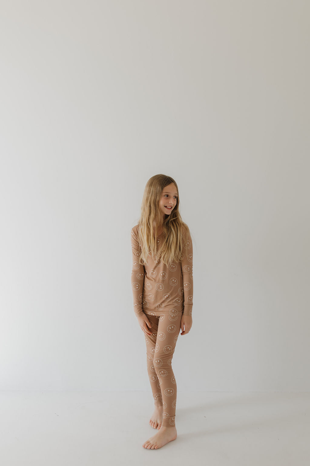 A young girl with long blonde hair stands against a plain light background, wearing breathable, hypoallergenic "Bamboo Two Piece Pajamas | Sandstone Smile" from forever french baby—beige with white smiley faces. She looks to the side and smiles, combining comfort and style effortlessly.