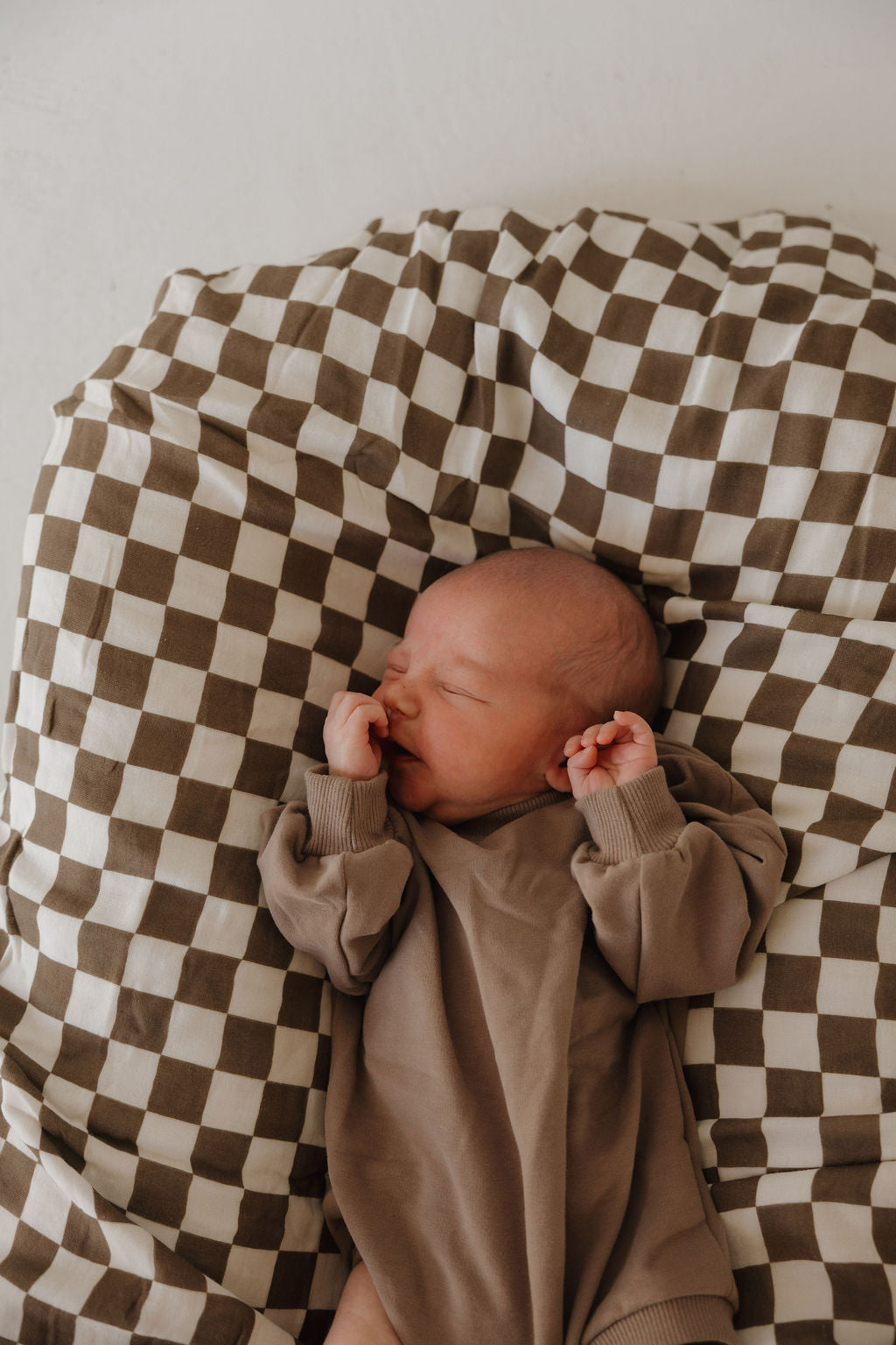 A newborn sleeps on a brown and white checkered blanket, wearing the forever french baby Sweatshirt Romper | Hot Cocoa with hands near their face.