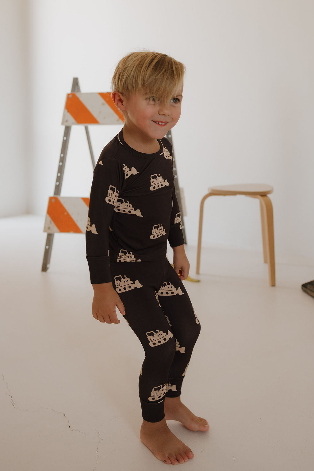 A young child with short blond hair wears the forever french baby Child Sleepy Time Set | Bulldozer—black breathable pajamas featuring construction vehicles—while standing barefoot in a minimal room with a wooden stool and caution-striped ladder.