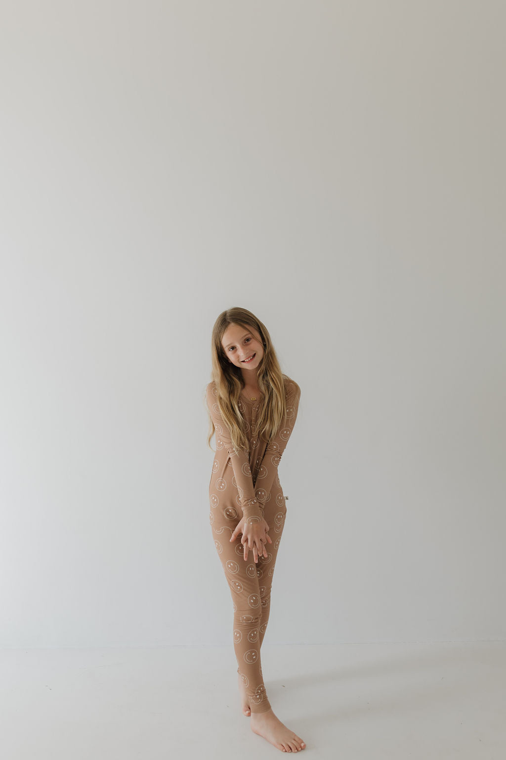 A young girl with long hair stands barefoot against a plain white background, wearing the "Bamboo Two Piece Pajamas | Sandstone Smile" by forever french baby. The beige suit features a subtle smiley face pattern and offers breathable comfort, allowing her to gently smile with her hands clasped in front.