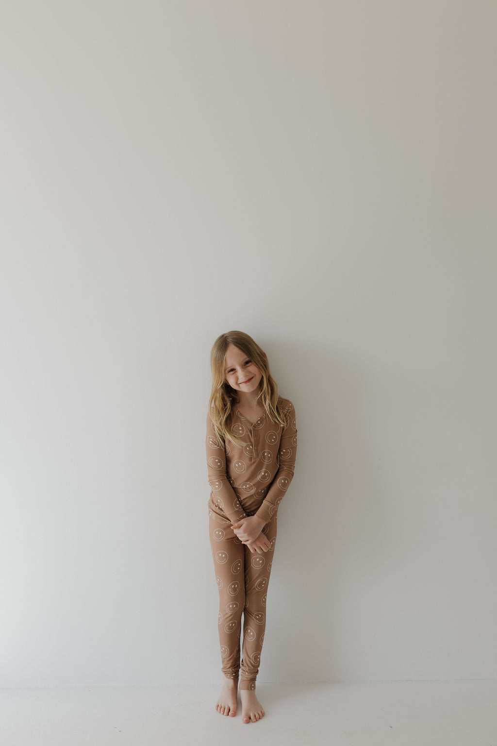A child with long hair, barefoot, and smiling gently stands against a white wall wearing forever french baby's hypo-allergenic Bamboo Two Piece Pajamas in Sandstone Smile.