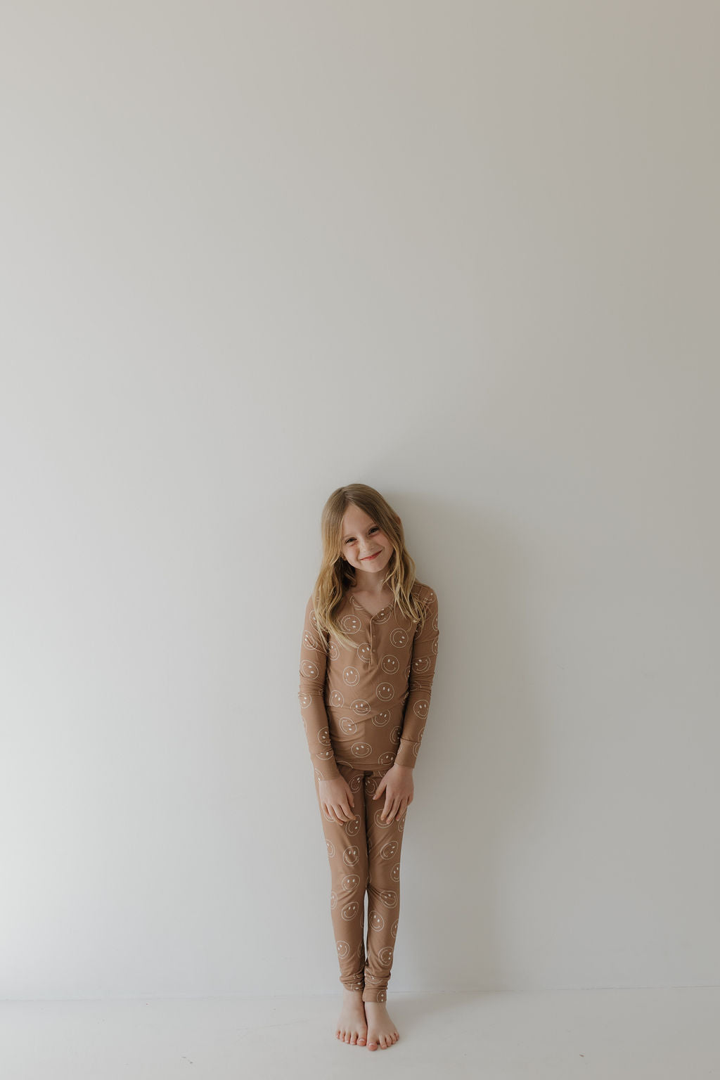 A young girl in forever french baby's Bamboo Two Piece Pajamas | Sandstone Smile stands barefoot against a white wall, smiling gently with hands clasped in front.