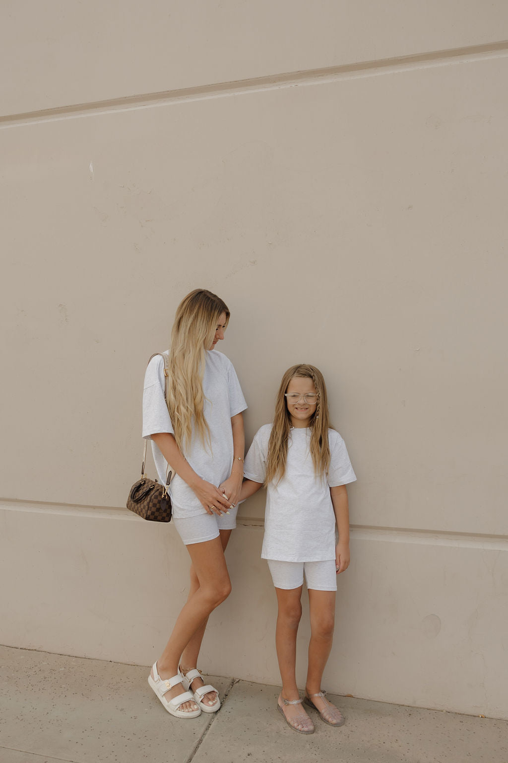A woman and a young girl stand against a beige wall in matching short sets; the girl wears the forever french baby Child Short Set | Heather, smiles, and has glasses. The woman has long blonde hair and carries a brown purse.