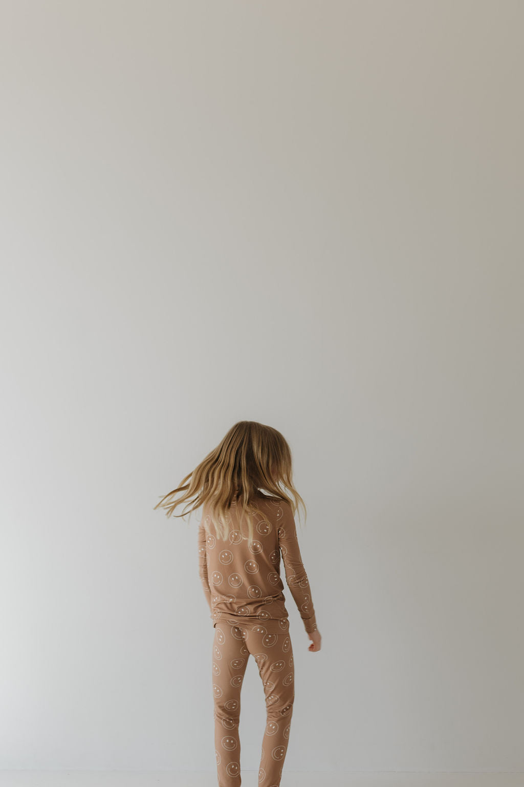 A child with long hair stands facing a plain light-colored wall, dressed in hypo-allergenic "Bamboo Two Piece Pajamas | Sandstone Smile" by forever french baby.