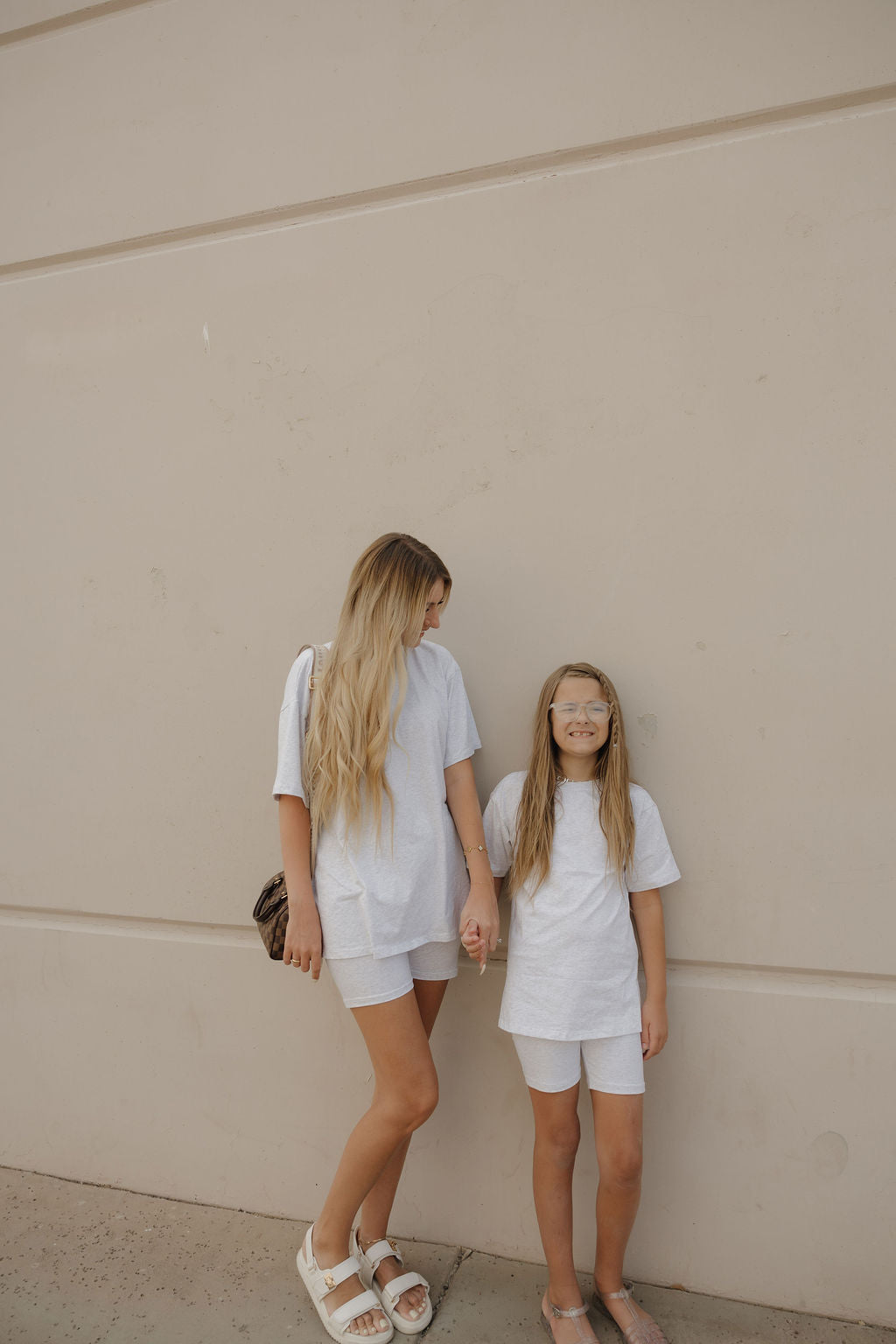 A woman and a young girl stand against a beige wall, smiling and holding hands in matching forever french baby Women's Short Set | Heather outfits with oversized tees. The woman has long blonde hair and sandals; the girl wears glasses.