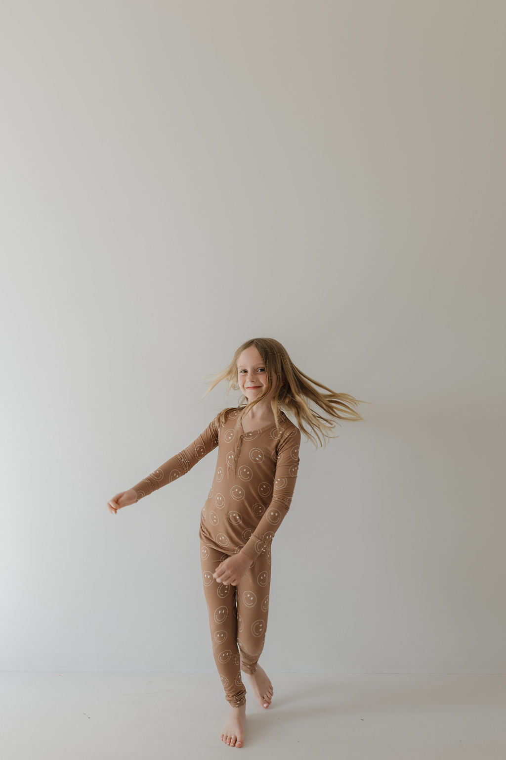 A young girl joyfully spins, her hair flying as she twirls in the breathable Bamboo Two Piece Pajamas | Sandstone Smile by forever french baby. The plain, light-colored wall enhances the simple and cheerful scene.