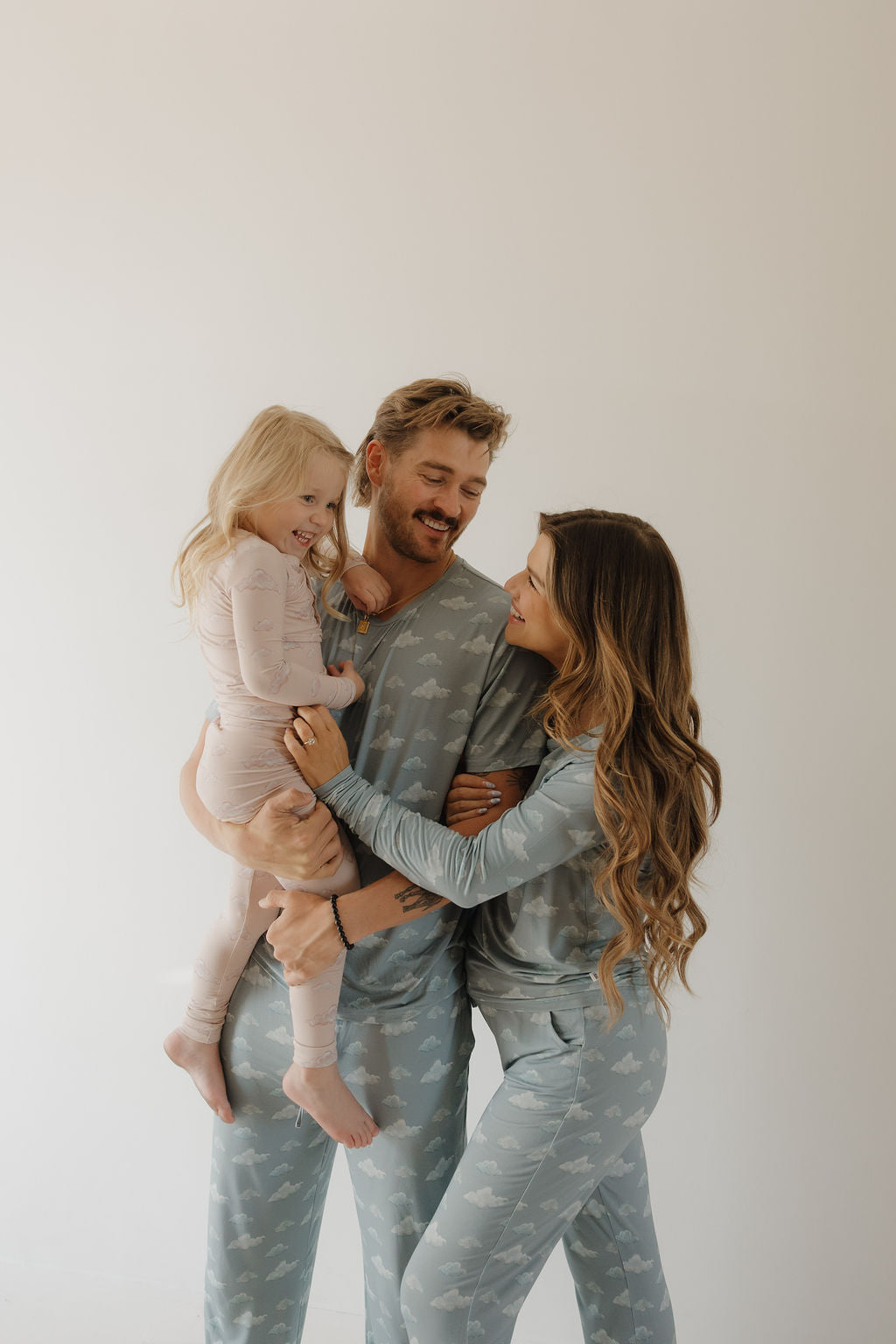 A woman in Kendy 3's Women's Bamboo Pajama | Duke, featuring a blue and white cloud pattern, smiles at a man while he holds a young girl in light pink pajamas. Their breathable fabric ensures comfort against a light background.