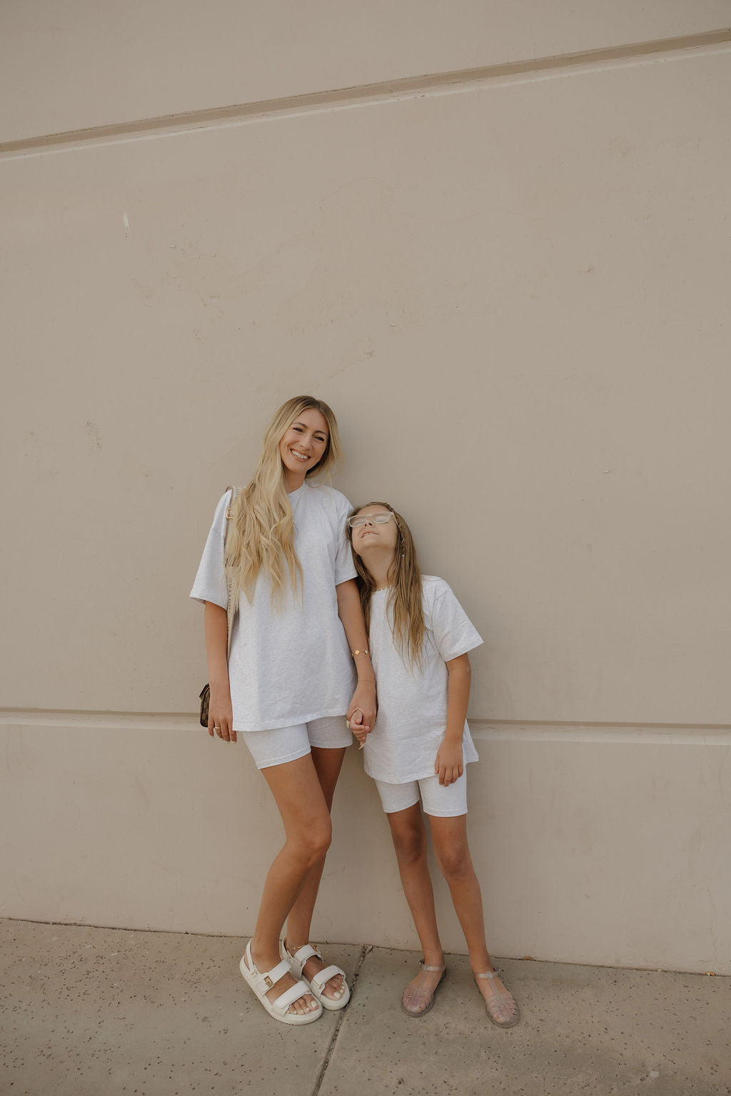 A woman and a young girl stand against a beige wall, both wearing matching forever french baby Women's Short Set | Heather. The woman smiles as the girl looks up at her, and they appear relaxed and happy together.