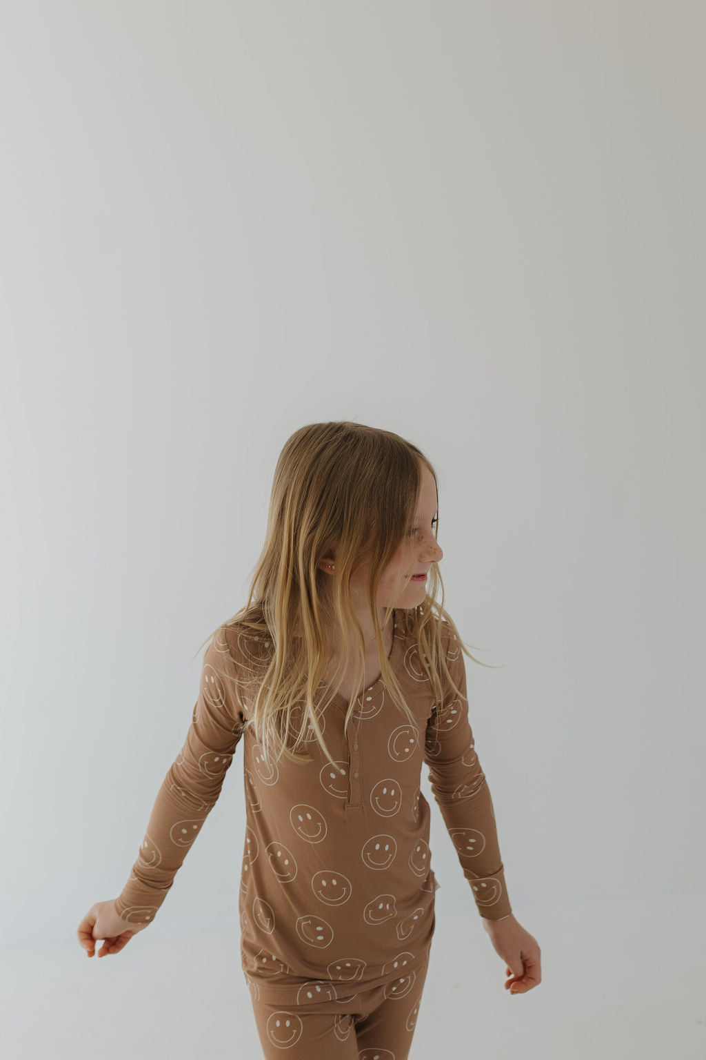 A young girl with long blond hair, wearing forever french baby's breathable Bamboo Two Piece Pajamas in Sandstone Smile, stands against a white background. She looks to her left with a relaxed posture and a slight smile.