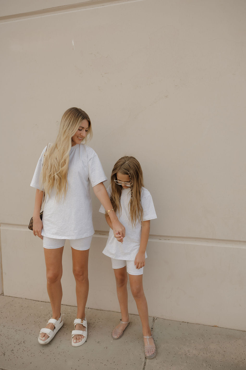 A woman and a young girl wear matching forever french baby Women's Short Set in Heather, standing against a beige wall. The woman smiles while holding the girl’s hand as she looks down.